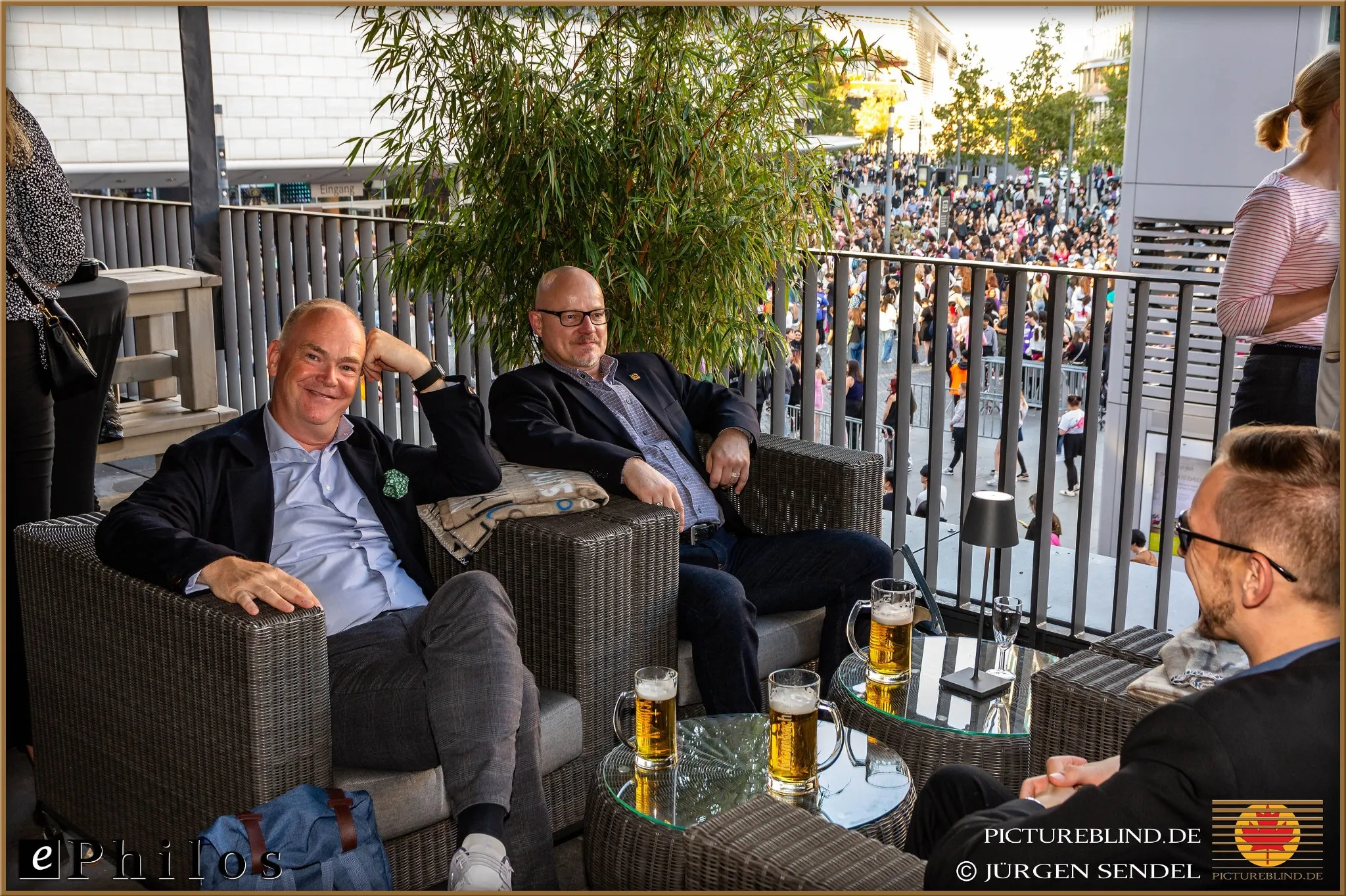 Geschäftstreffen von Ephilos: Drei Männer sitzen entspannt auf einer Terrasse mit Blick auf eine belebte Veranstaltung, genießen Bier und diskutieren über Softwarelösungen