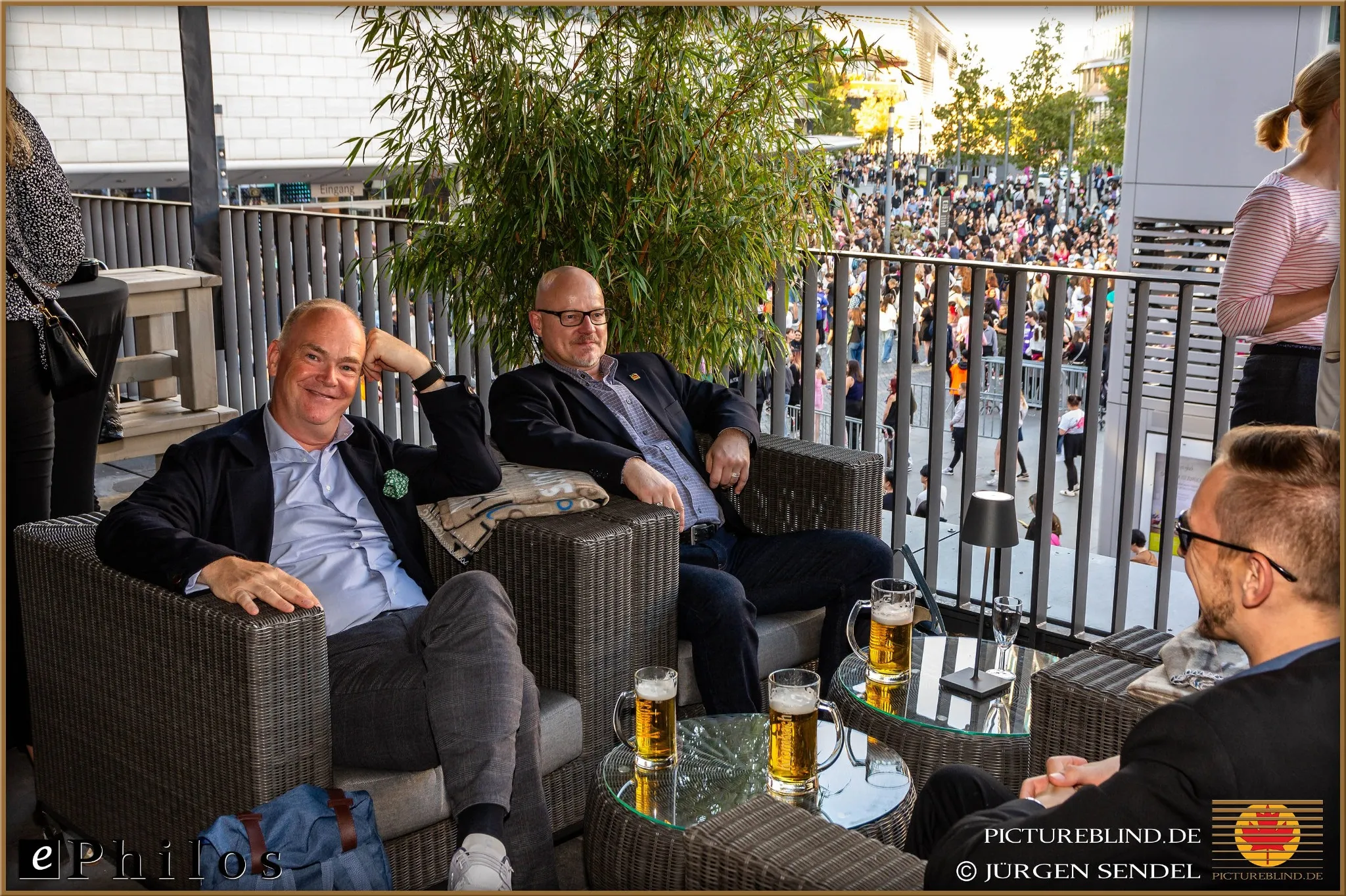 Ephilos business meeting: Three men sitting relaxed on a terrace overlooking a busy event, enjoying beer and discussing software solutions