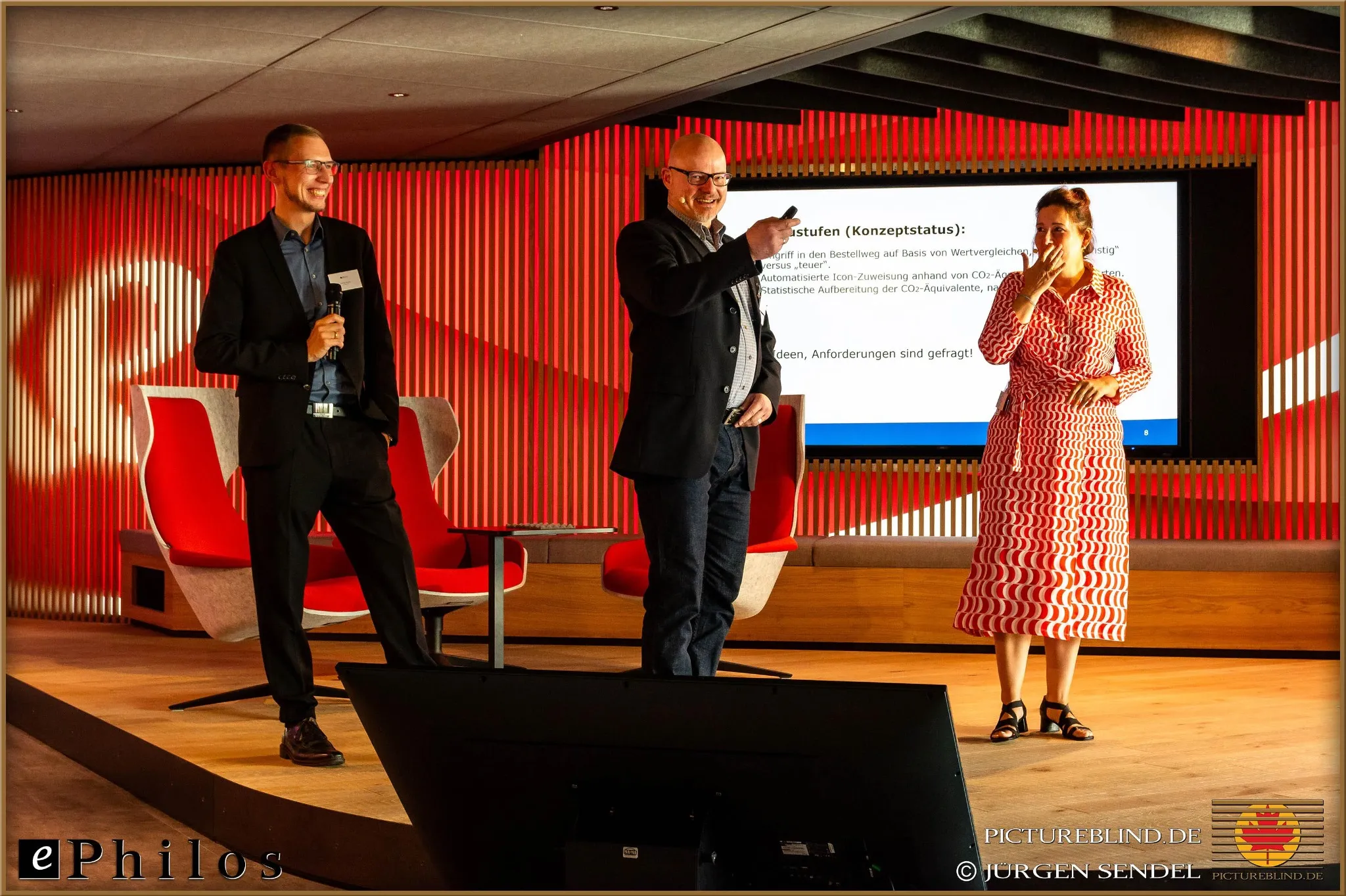 Three presenters on stage in warm lighting in front of a large screen during a business event