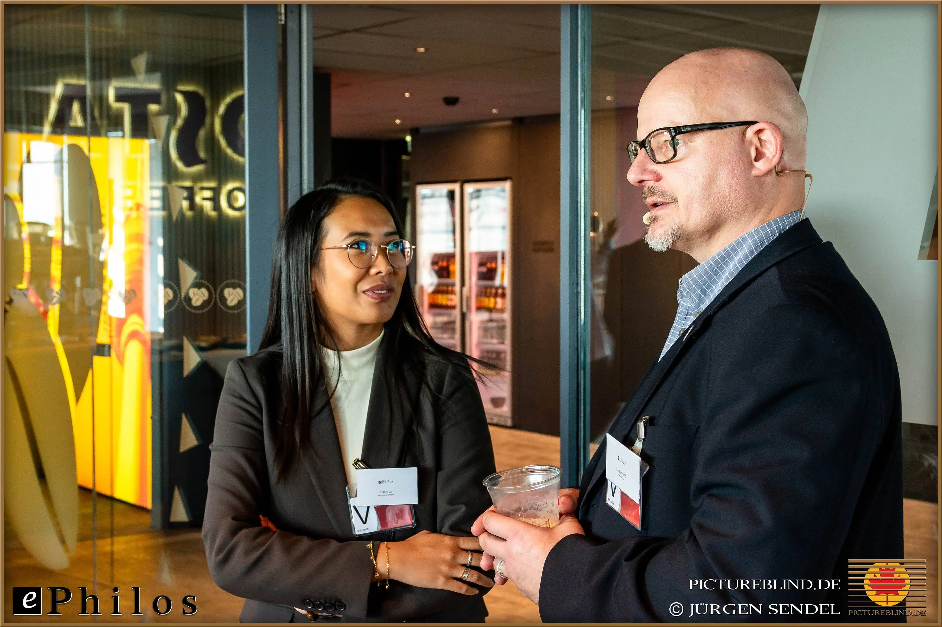 Two business participants talking during a break, in the background a modern break area with refrigerators and a bright sign