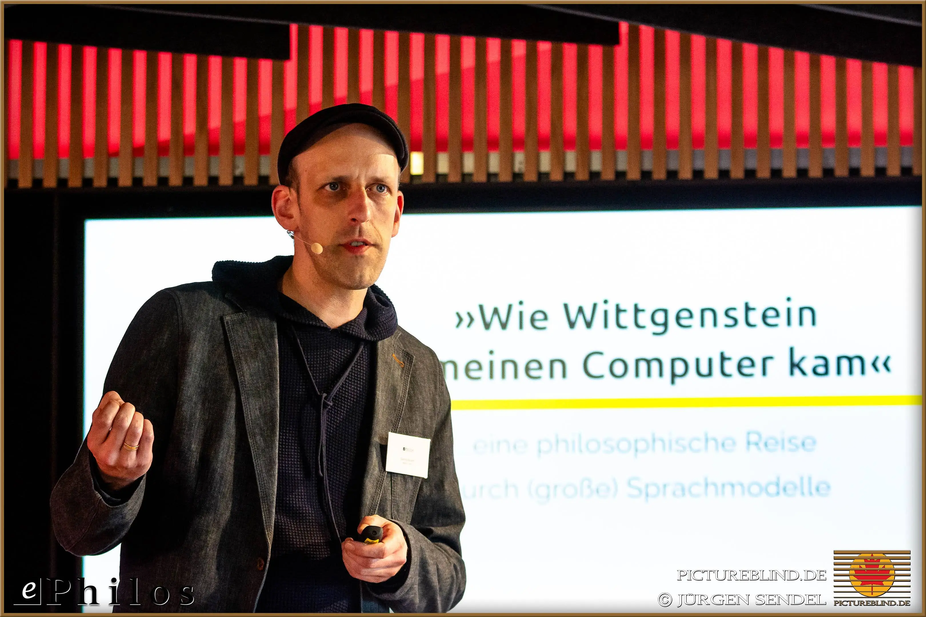 Presenter giving a talk titled 'How Wittgenstein Came to Have a Computer' in front of a screen during a business event