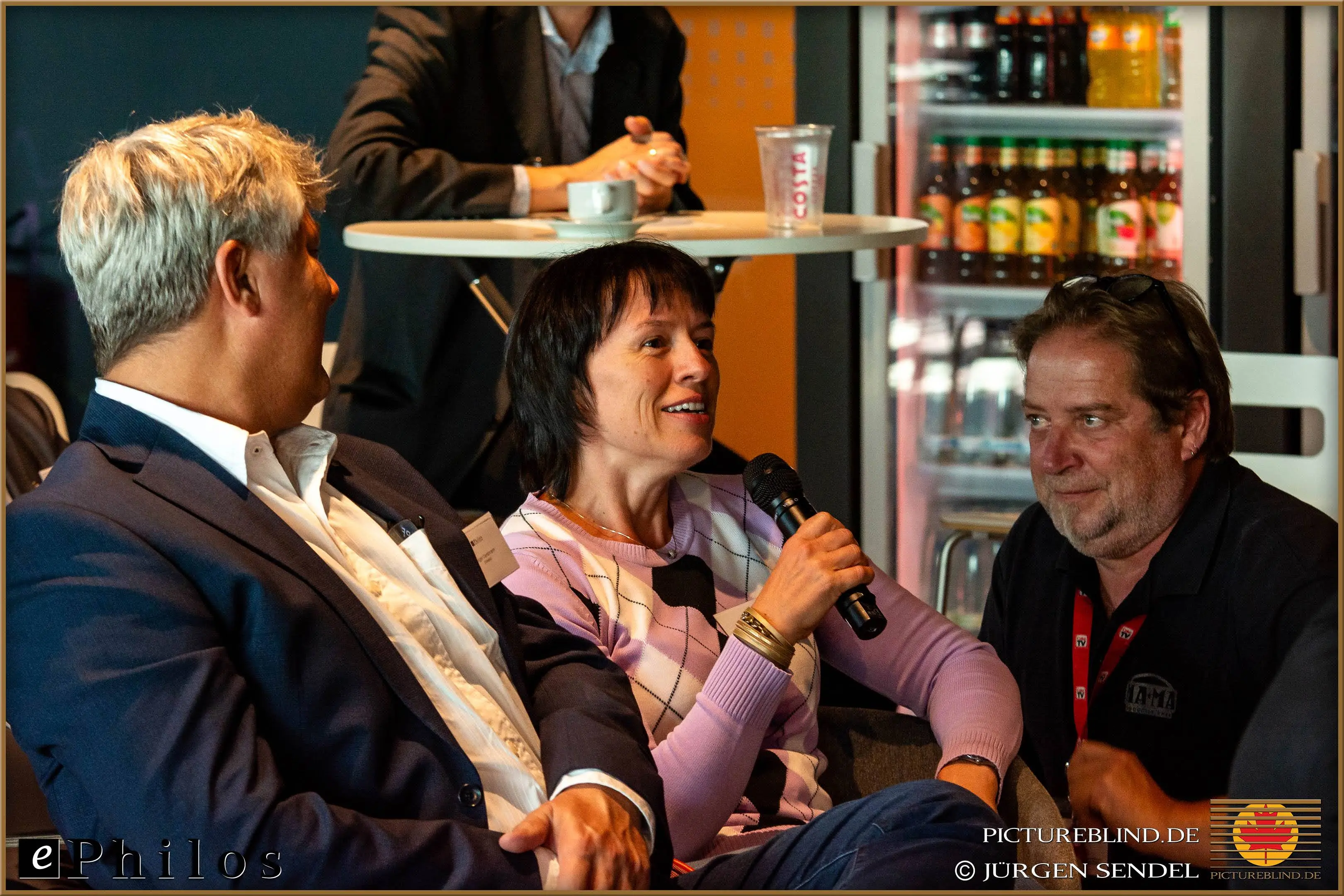 Teilnehmerin spricht mit einem Mikrofon, während sie in einer Lounge sitzt, umgeben von anderen Teilnehmern und Getränken im Hintergrund