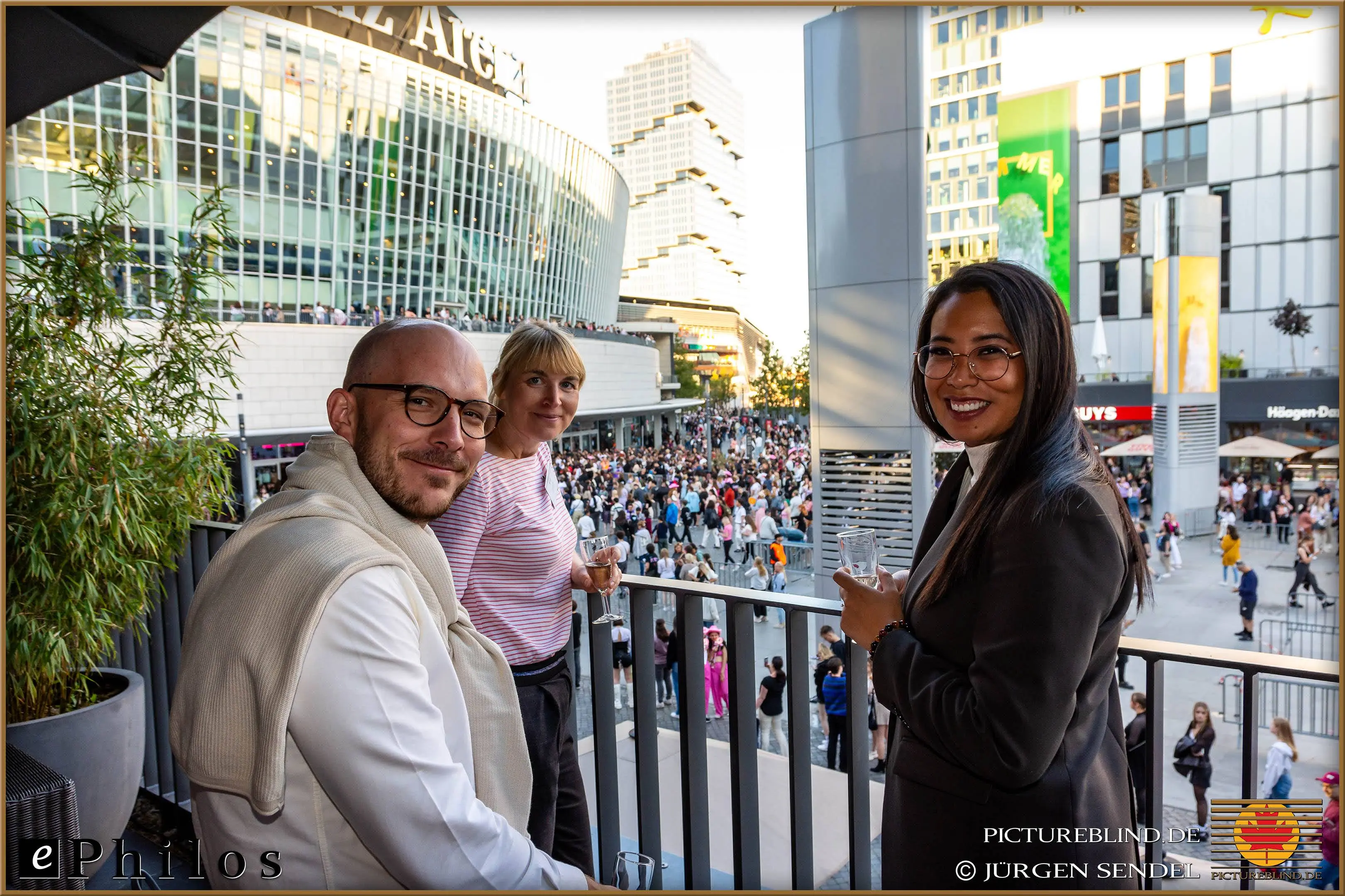 Drei Teilnehmer genießen die Aussicht von einem Balkon mit Blick auf eine belebte Veranstaltung am Mercedes-Benz-Platz