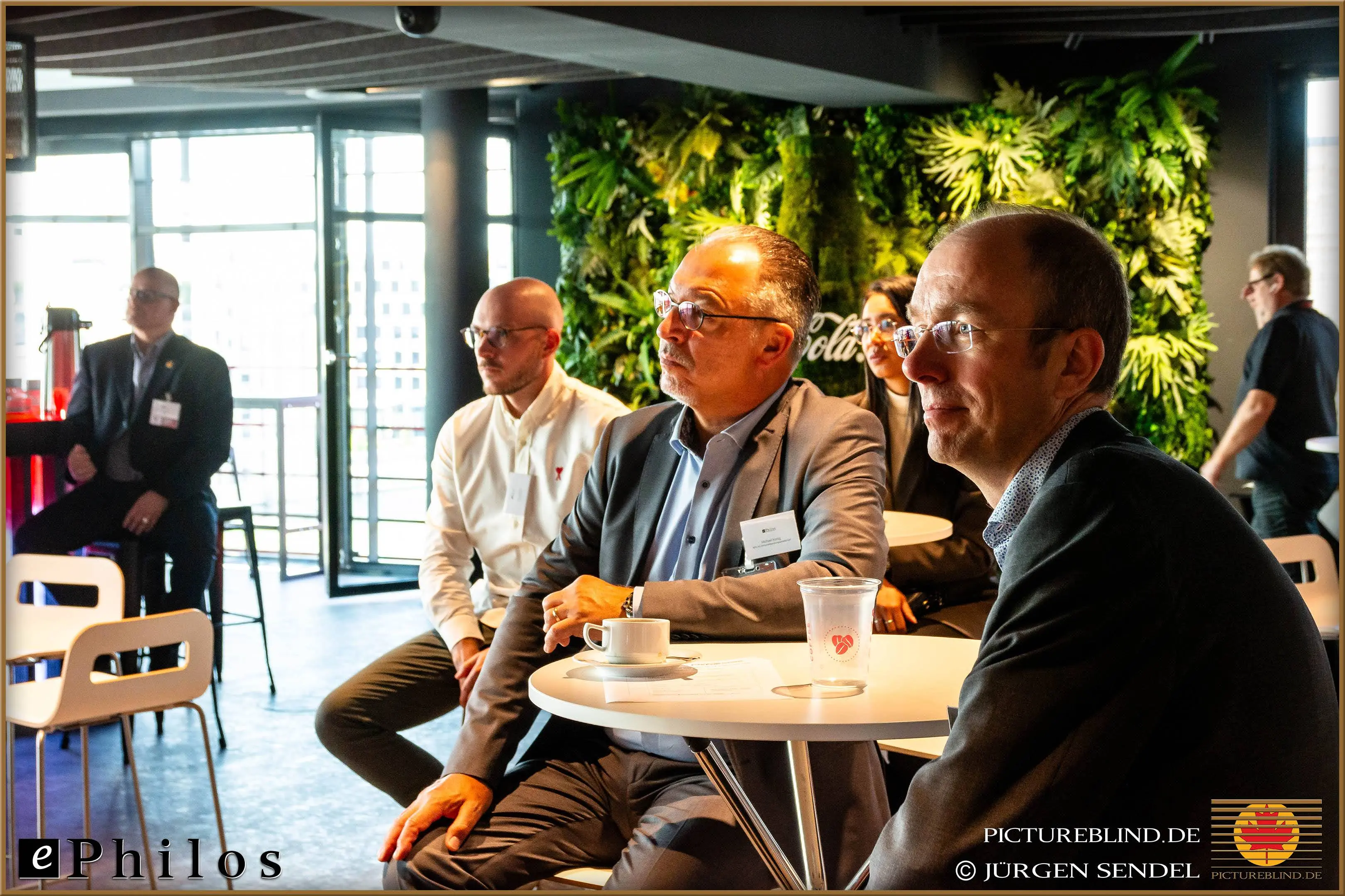 Participants of a business event sitting at tables and listening attentively, with a green plant wall and a bright conference room in the background